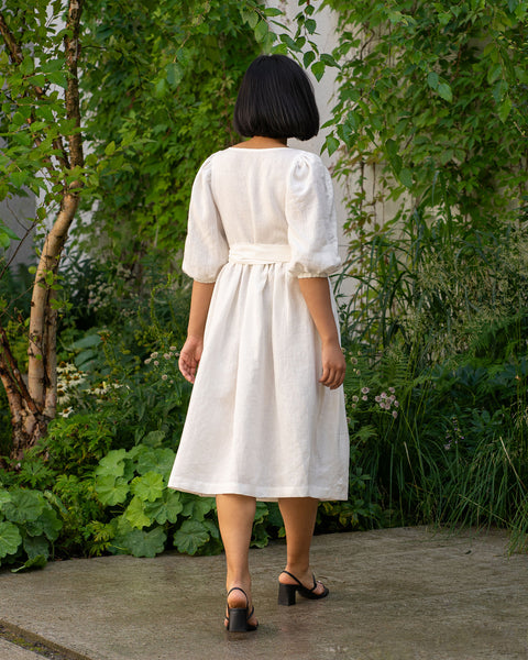From the back, a person with short, dark hair is walking away on a paved surface surrounded by greenery. They are wearing a pure white dress with elbow-length puff sleeves and a tied waist, along with dark-colored heeled sandals.