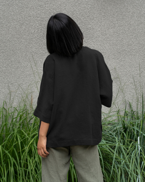 Woman with short dark hair wearing a pure black linen jacket and stone green menique pants, viewed from the back.