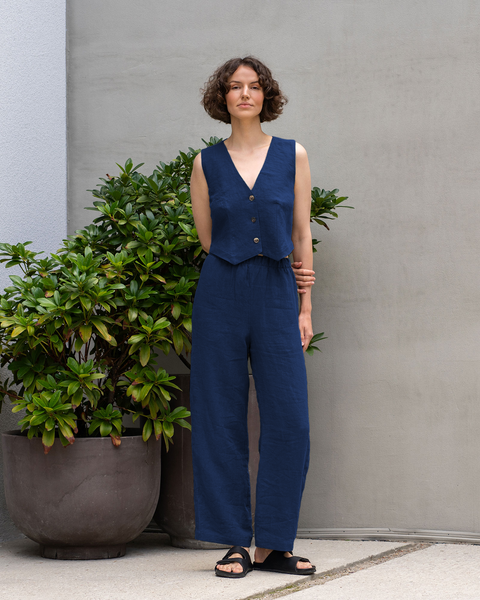 A full-body shot of a woman wearing a menique storm blue linen two-piece outfit consisting of a vest and wide-leg pants. She is standing next to a large green plant in a brown pot, with a textured grey wall in the background.