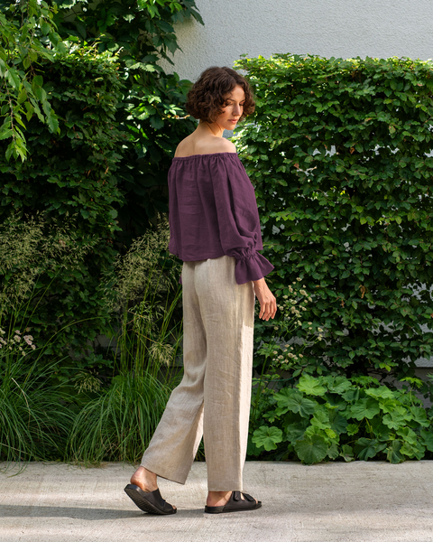 A woman in an off-the-shoulder shadow purple linen menique top and beige pants walks on a stone path near greenery.