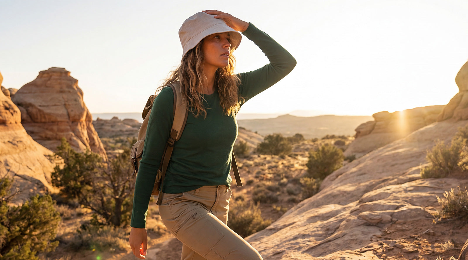 A woman wearing a light-colored hat and a green top looking out over a rocky, canyon-like landscape during golden hour.