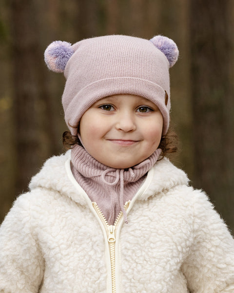 A young child with light brown curly hair and a subtle smile, looks directly at the viewer. They are wearing a light pink knit hat with two lighter pink pom-pom ears, a matching pink turtleneck or neck warmer, and a cream-colored sherpa jacket with a zipper. The background is a blurred forest.