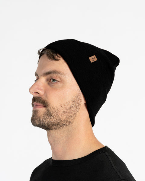 Man in side profile wearing a black merino wool knit beanie with small leather logo patch, studio background.