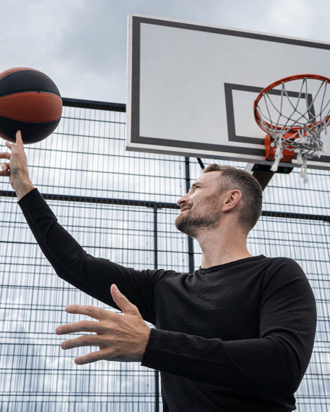 A man standing on an outdoor basketball court, looking up as he balances and spins an orange and black basketball on his index finger. He is wearing a black long-sleeved top. A basketball hoop and a tall wire fence are visible in the background against a cloudy sky.