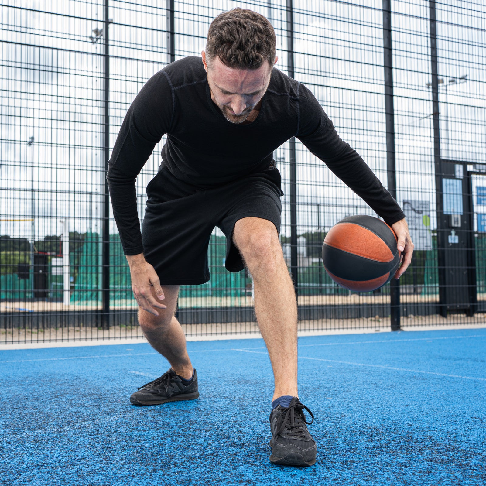 A smiling man sitting casually on a bright blue outdoor sports court, leaning back on one hand. He is wearing a black long-sleeved athletic top and black shorts, with an orange and black basketball resting on the ground next to his leg.