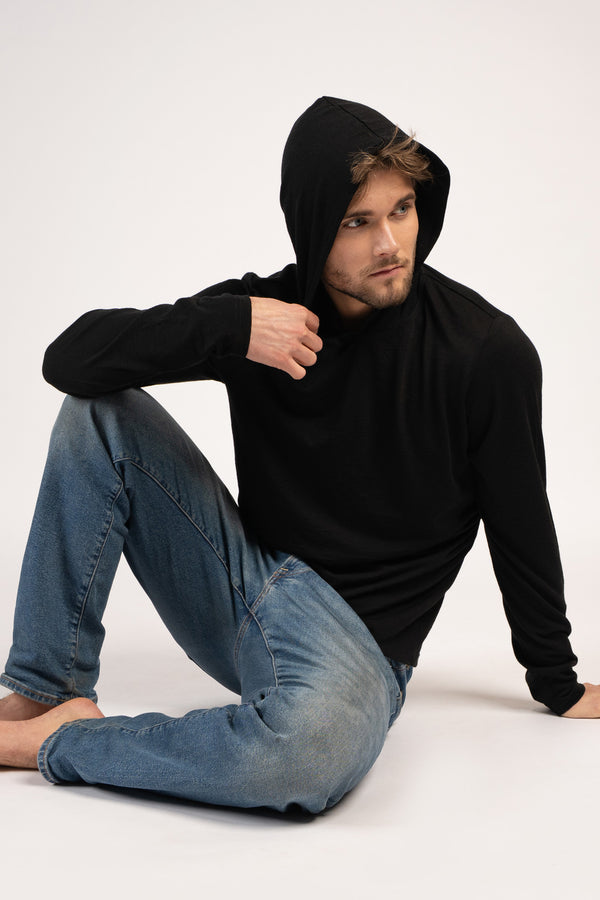 A barefoot man sits on the floor wearing blue jeans and a black long-sleeved hooded shirt with the hood up. He is looking off to the right with one hand resting near his collar and the other on the floor.