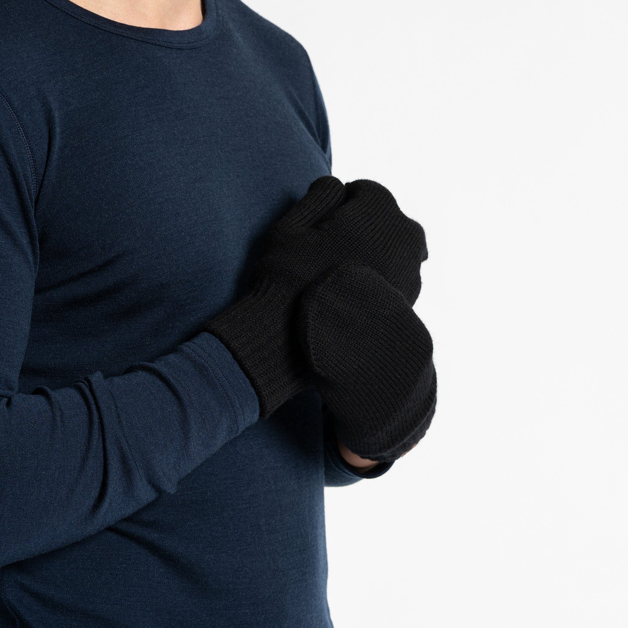 A person wearing a dark blue long-sleeved shirt and black menique mittens with a flap that can cover the fingers, turning them into mittens. The person is holding their hands together, and the flap on one glove is pulled back, exposing the fingers, while the flap on the other glove covers the fingers.