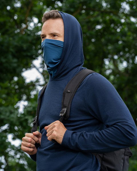A man standing outdoors against a backdrop of green trees. He is wearing a navy blue long-sleeved hoodie with the hood pulled up, and a denim-blue neck gaiter pulled over his nose and mouth. He is holding the shoulder straps of his black backpack and looking off to the side.