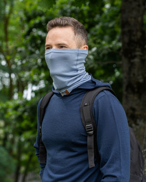 A man standing outdoors with green trees in the background. He is wearing a navy blue long-sleeved hoodie and a black backpack. A light dusty-blue neck gaiter is pulled up over his nose and mouth, and he is looking off to the side.