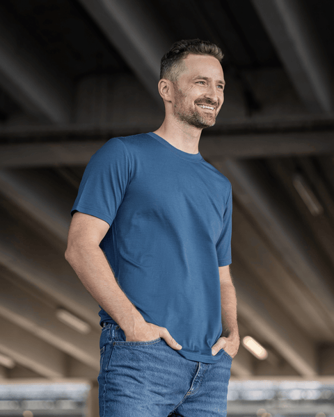 A man with short hair and a beard wearing a plain blue t-shirt and blue jeans, standing with his hands in his pockets under a concrete overpass.