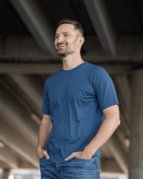 A man in a blue t-shirt and jeans standing under a concrete structure, smiling and looking off to the side with his hands resting in his front pockets.