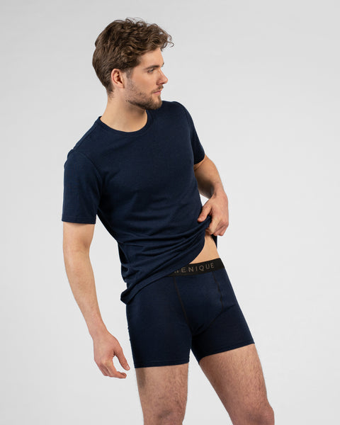 A studio portrait of a man looking to the right while lifting the hem of his navy blue short-sleeved t-shirt. He pairs it with matching navy blue short boxer briefs featuring a black logo waistband.