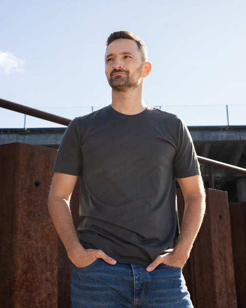 A man stands outdoors on a sunny day, wearing a charcoal grey short-sleeved t-shirt and blue jeans with his hands tucked into his front pockets. He is looking slightly off to the side against a backdrop of rusted steel beams and a concrete overpass.
