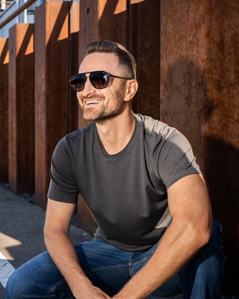 A smiling man wearing a charcoal grey short-sleeved t-shirt, blue jeans, and sunglasses crouches outdoors in bright sunlight. He is looking off to the left, positioned in front of a row of tall, rusted steel pillars.