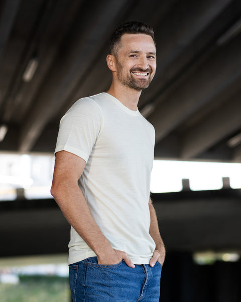 A smiling man standing outdoors under a concrete structure, looking directly at the camera. He is wearing a natural, off-white short-sleeve crew neck t-shirt and blue jeans, with his hands resting in his front pockets.