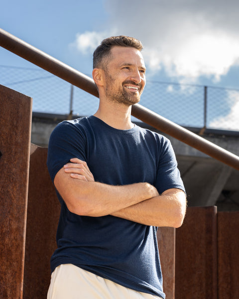 A smiling man standing outdoors on a sunny day with his arms crossed, looking off into the distance. He is standing near a rusty metal structure and wearing a short-sleeve navy blue crew neck t-shirt and light-colored shorts.
