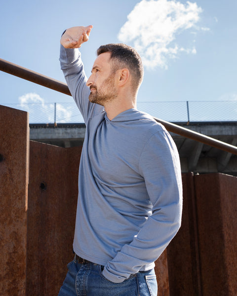 A man standing outdoors near a rusted metal structure on a sunny day. He is wearing a dusty blue hoodie and blue jeans, holding one hand up to shield his eyes from the sun as he looks off into the distance.
