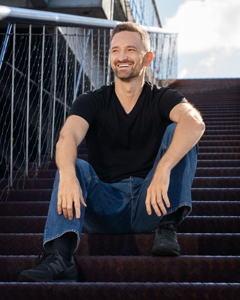 A smiling man sitting on outdoor metal stairs. He is wearing a short-sleeve black V-neck t-shirt, loose blue jeans, and black sneakers, resting his hands casually on his knees.