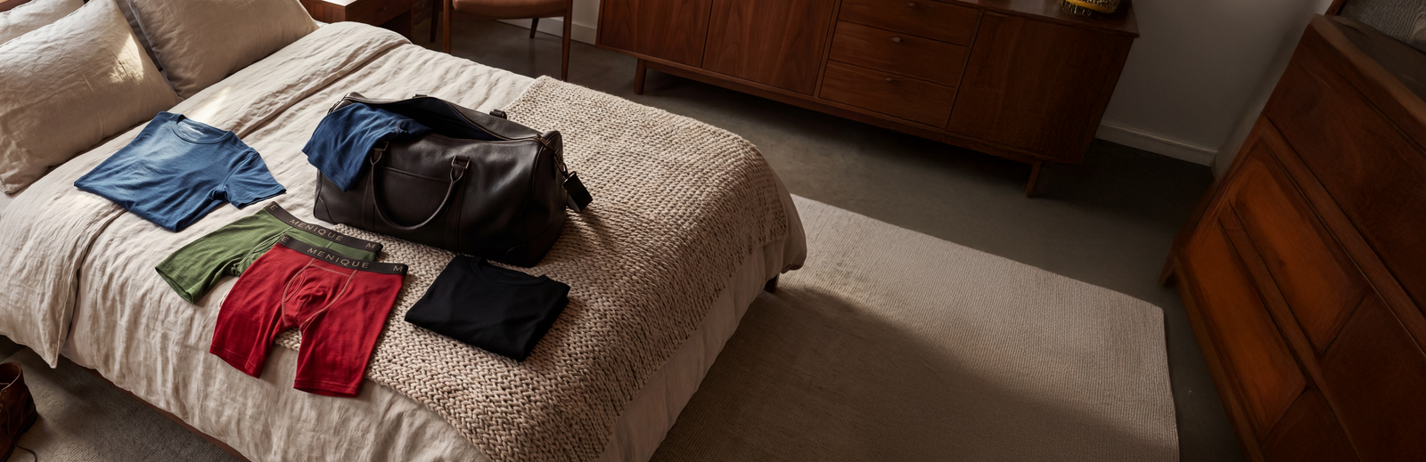 A bedroom scene showing an open, dark brown leather duffel bag resting on a light-colored textured bedspread. Neatly laid out around the bag are packing items including a folded blue t-shirt, a folded black shirt, and two pairs of Menique brand boxer briefs in olive green and deep red. Another blue garment is partially tucked into the open bag.