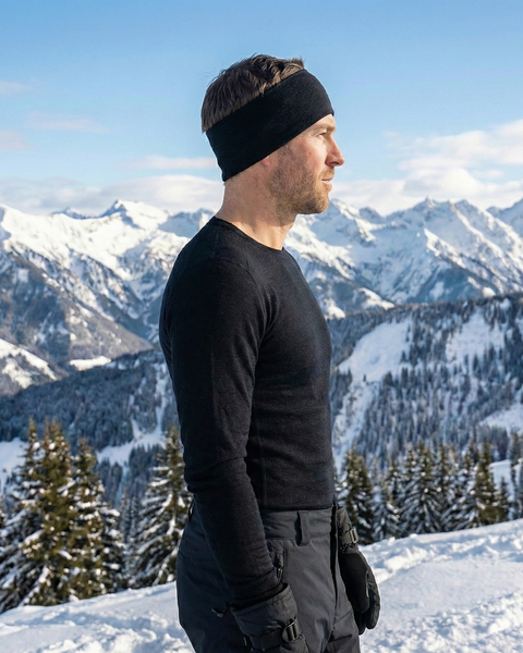 Man wearing a black merino wool headband and long-sleeve base layer, standing on a snow-covered mountain with alpine scenery.