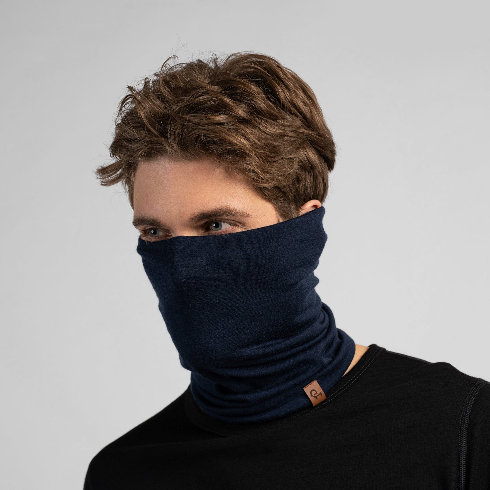 A dark navy blue tubular neck gaiter displayed against a solid white background. The soft fabric is styled with natural-looking folds and slouching, and a small, rectangular brown leather logo tag is attached near the bottom edge.