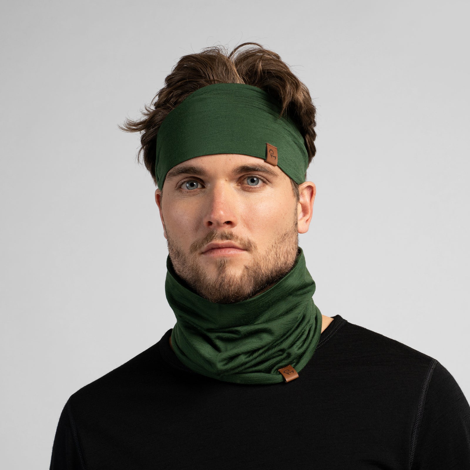 A studio portrait of a man looking off to the left. He is wearing a black long-sleeved shirt, a dark green headband, and a matching dark green neck gaiter pulled up to cover his mouth and nose.