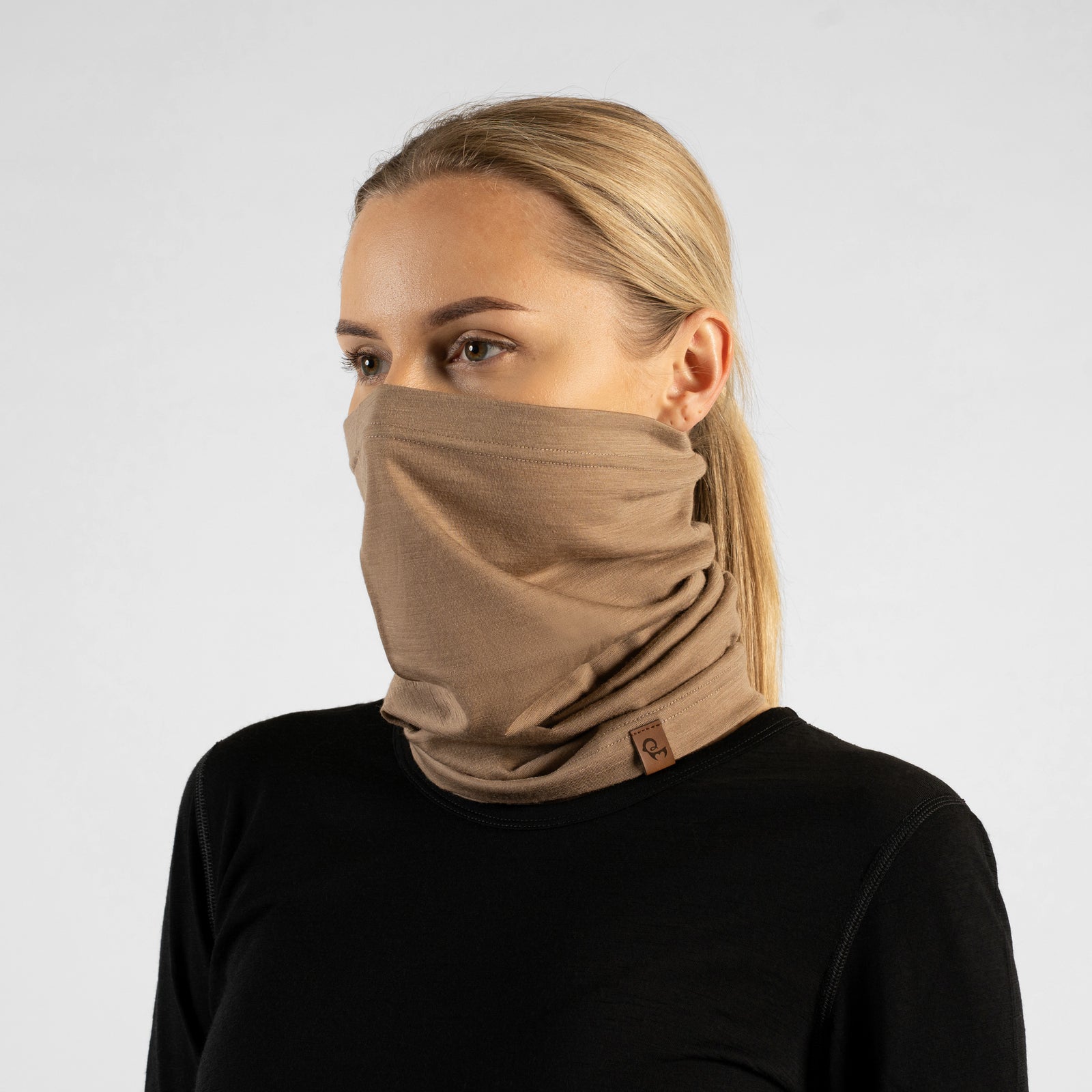 A woman standing outdoors wearing a black long-sleeved top. She has a beige neck gaiter pulled up over her nose and mouth. She is looking at the camera, smiling with her eyes, with one hand on her hip and the other hand resting behind her head, with a background of blurred trees and grass.