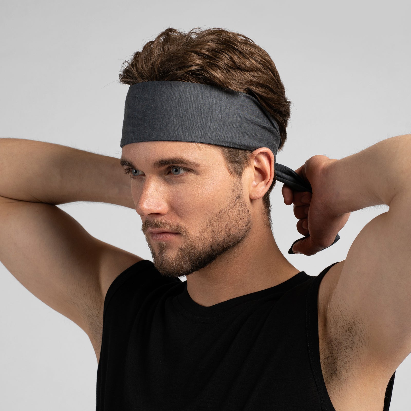 A studio portrait of a man looking thoughtfully off to the left side of the frame. He is wearing a medium grey tie-back headband, with the tied ends hanging down behind his neck. He is wearing a black sleeveless top against a solid light grey background.
