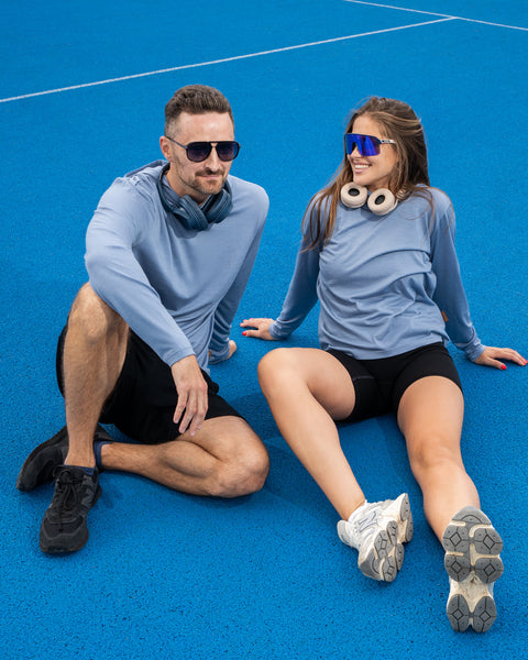 A man and a woman wearing matching light blue long-sleeved shirts, black shorts, and sunglasses, sitting casually on a bright blue sports court.