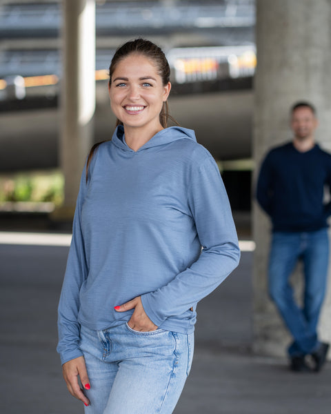 A smiling woman wearing a light blue hoodie and jeans standing under a concrete overpass, with a man standing in the blurred background.