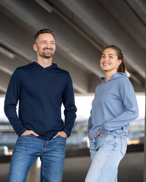 A man and a woman standing side-by-side outdoors under a concrete structure, both smiling and looking off into the distance. The man is wearing a navy blue hoodie and blue jeans, while the woman is wearing a dusty blue hoodie and light blue jeans. Both have their hands casually resting in their pockets.