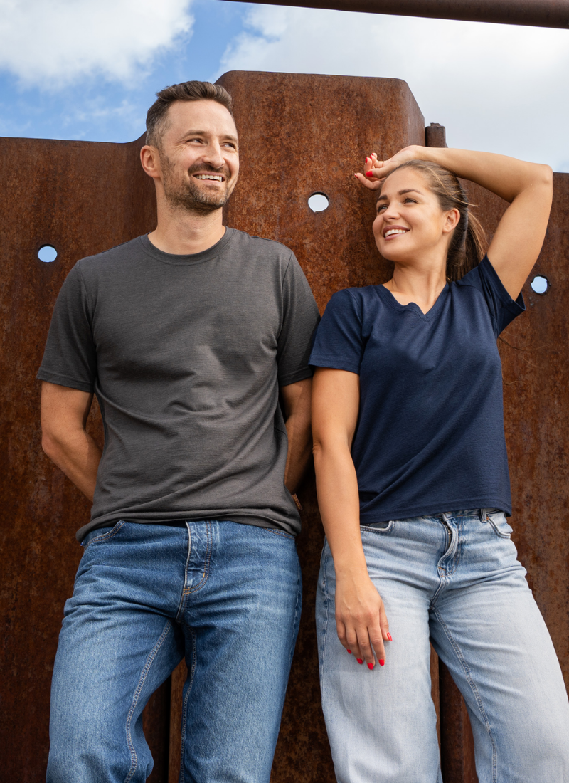 A smiling man and woman standing outdoors against a rusty, corrugated metal wall, modeling plain t-shirts. The man wears a dark grey t-shirt and blue jeans with his hands behind his back, while the woman wears a navy blue t-shirt and light blue jeans, resting one hand behind her head.