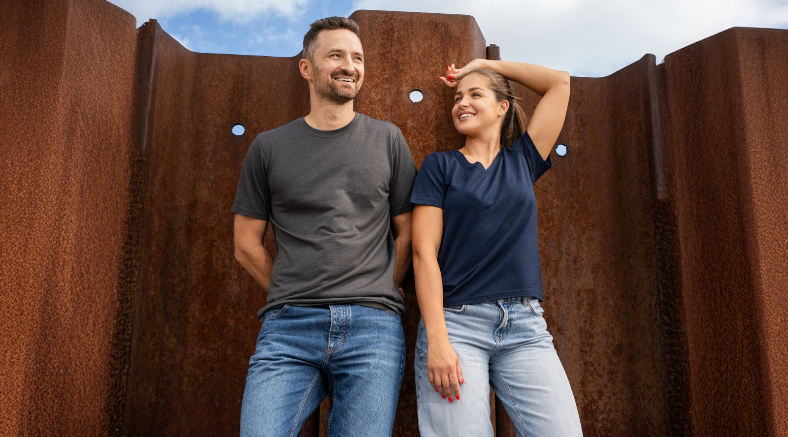 A smiling man and woman standing outdoors against a rusty, corrugated metal wall, modeling plain t-shirts. The man wears a dark grey t-shirt and blue jeans with his hands behind his back, while the woman wears a navy blue t-shirt and light blue jeans, resting one hand behind her head.