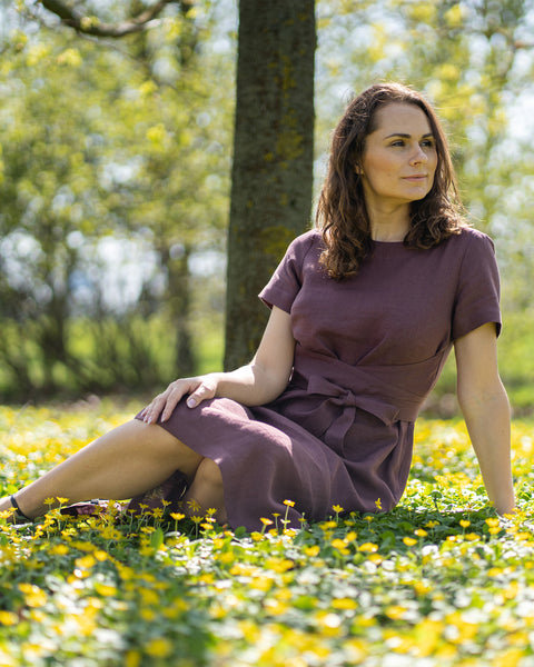 A woman with brown hair, wearing a short-sleeved purple dress, sits outdoors on the grass among small yellow flowers.
