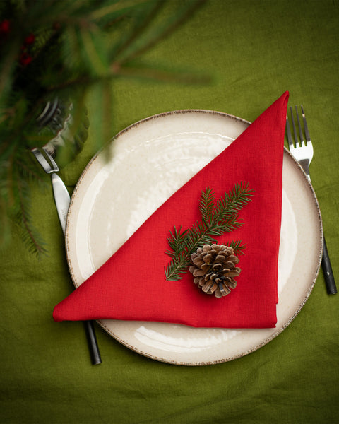 A top-down view of a Christmas place setting with a cream-colored plate on a green tablecloth. A red linen napkin is folded into a triangle and decorated with a pinecone and fir sprig. Silverware is placed on either side, and evergreen branches appear at the top edge of the image.