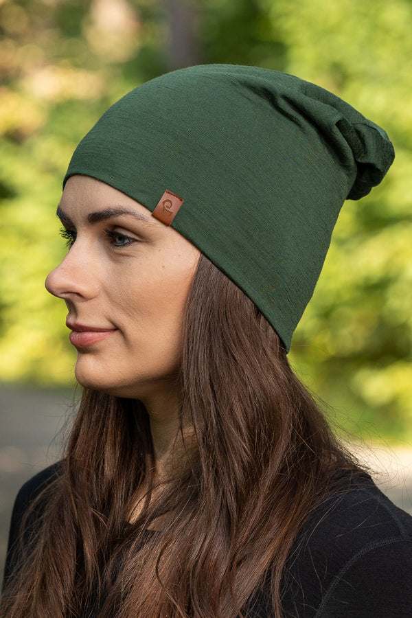 The picture features a side profile head-shot of a woman wearing the dark green unisex beanie. It is made from merino wool, has a small logo on the side at the front. Behind her is a blurred view of trees .