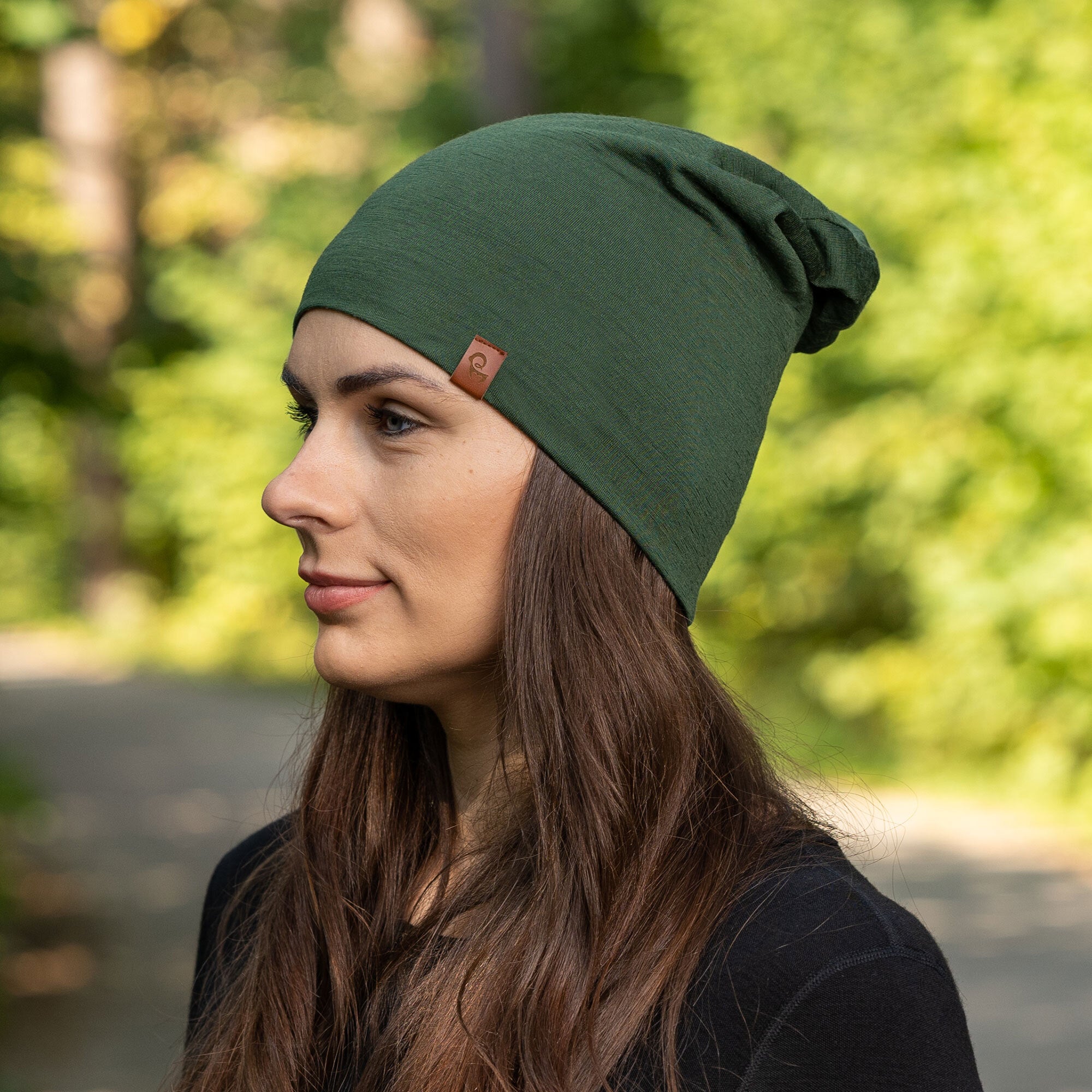 The picture features a side profile head-shot of a woman wearing the dark green unisex beanie. It is made from merino wool, has a small logo on the side at the front. Behind her is a blurred view of trees .