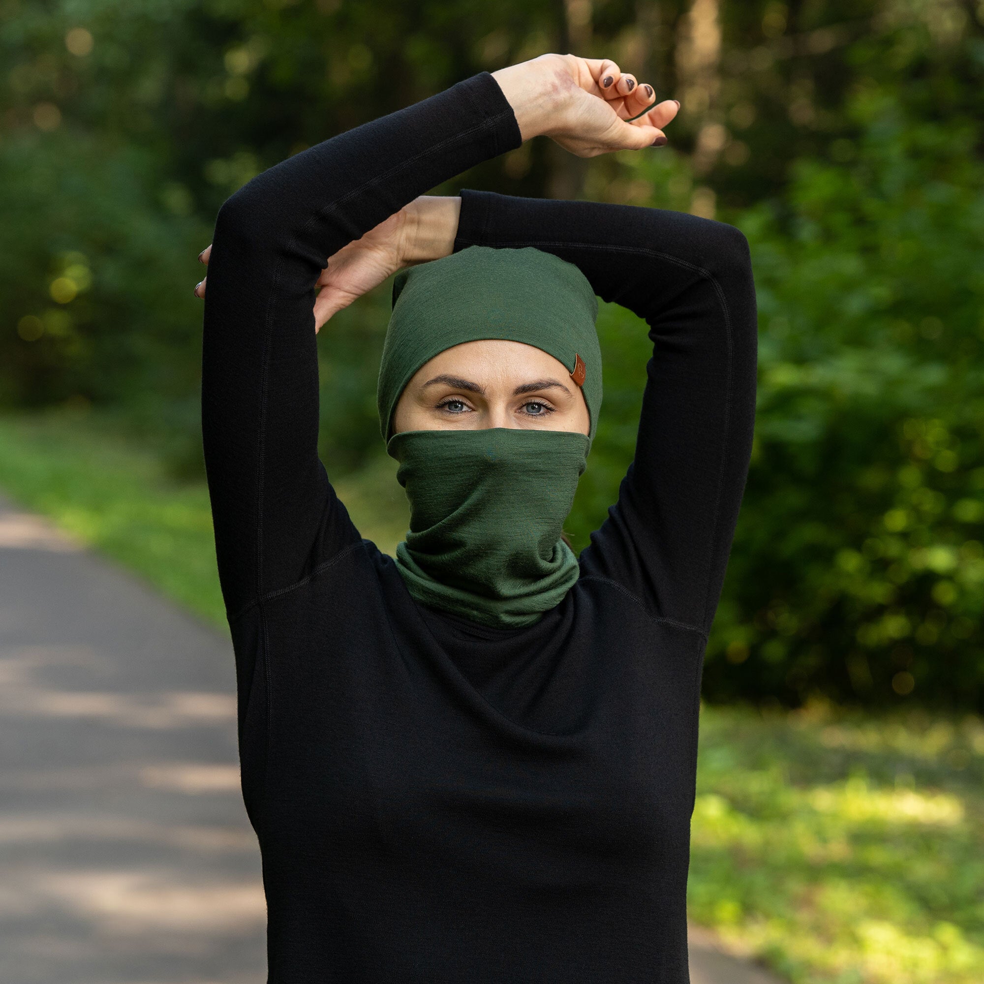 The picture features a man and a woman wearing the unisex dark green merino gaiter and beanie set. They are in a forest. The woman is pulling the man's gaiter to remove it. Both of them are smiling. 