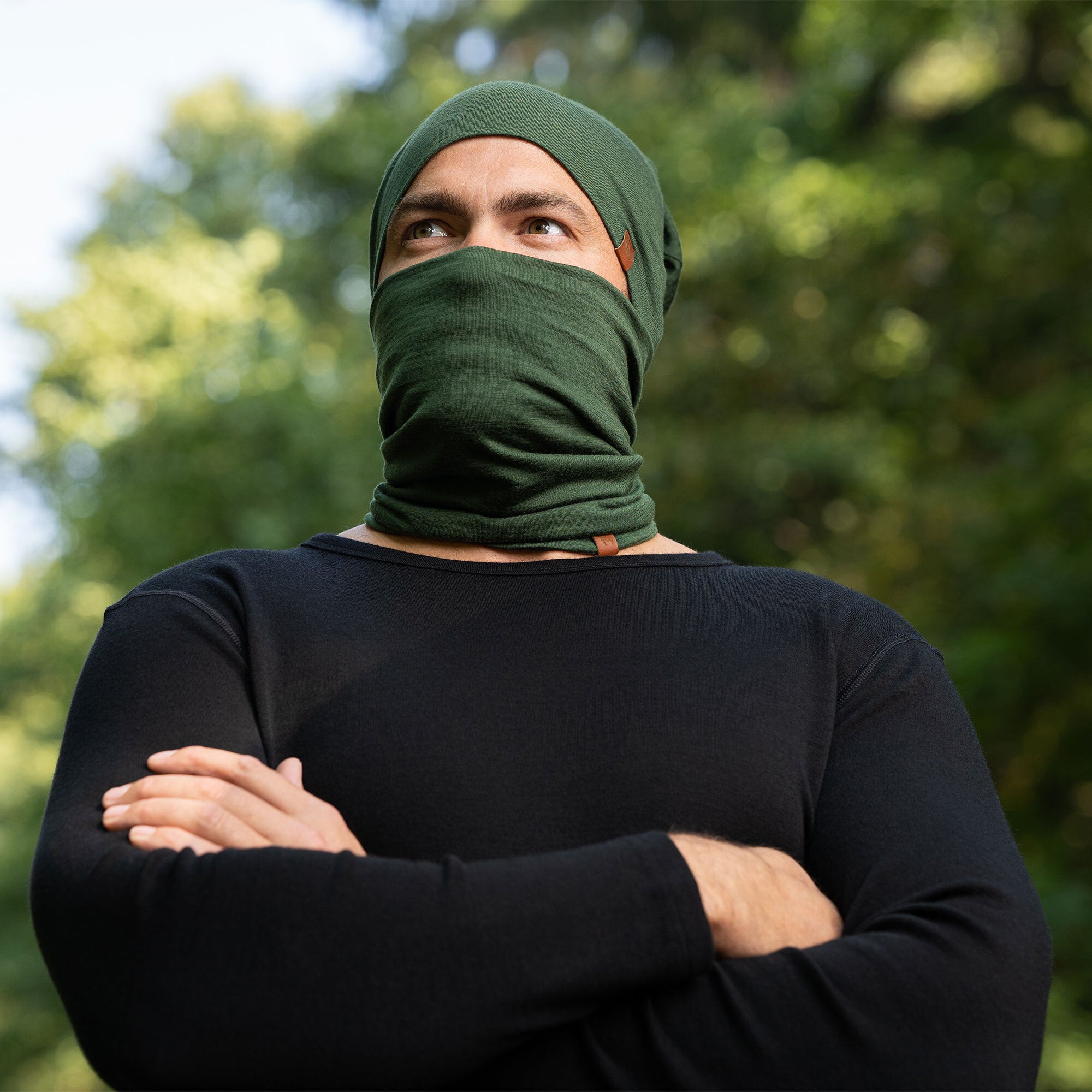 The picture features a man and a woman wearing the unisex dark green merino gaiter and beanie set. They are in a forest. The woman is pulling the man's gaiter to remove it. Both of them are smiling. 