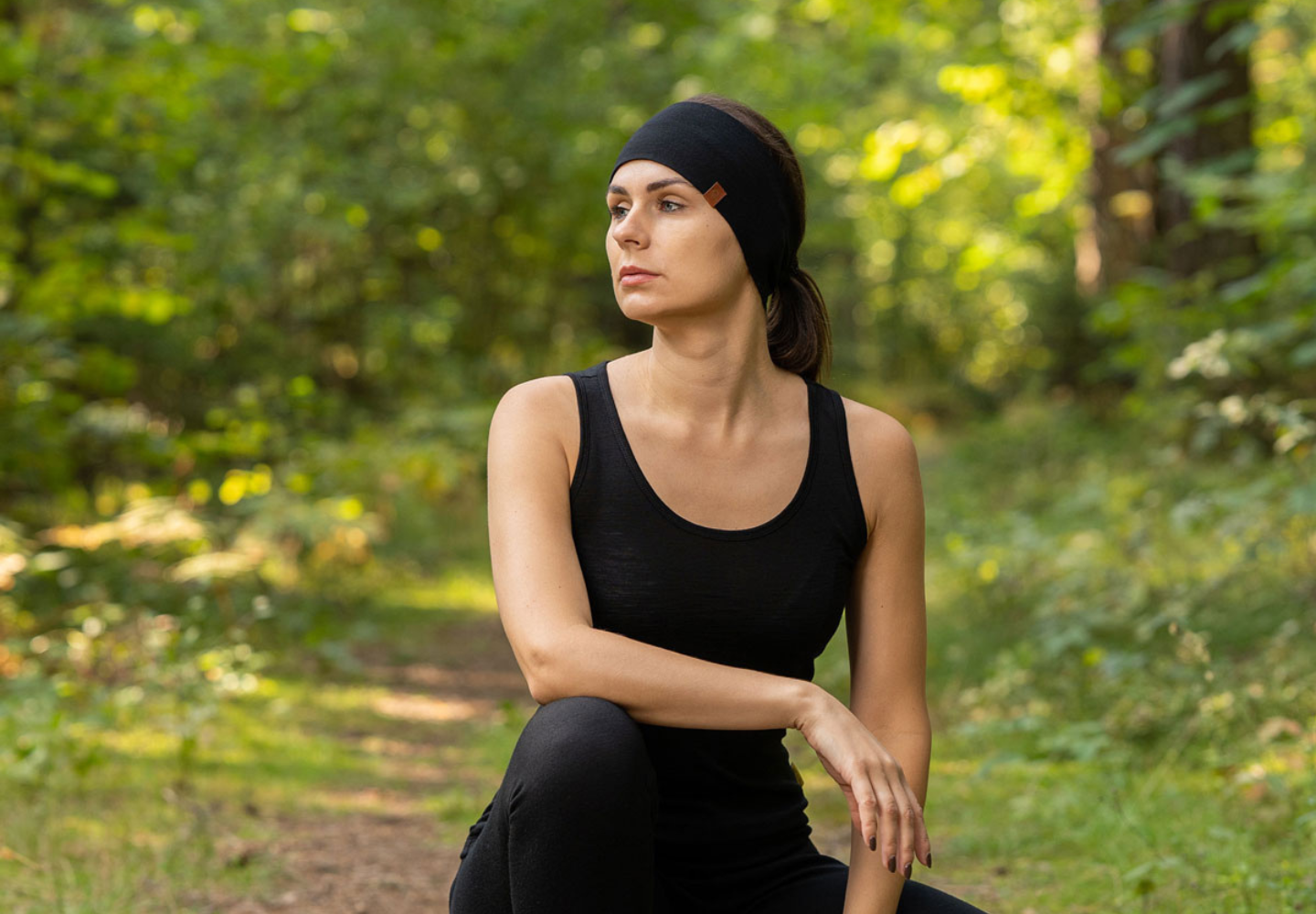 A woman wearing a black merino wool tank top and headband from menique kneels on a forest trail, surrounded by lush greenery and dappled sunlight.