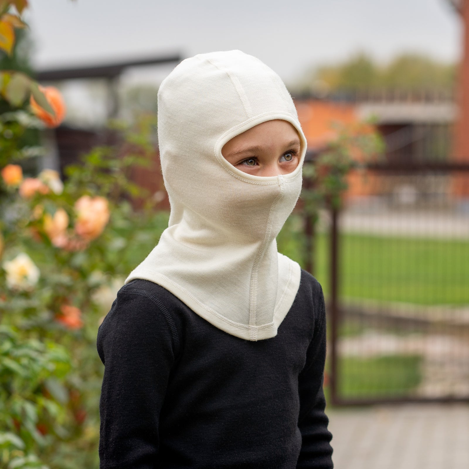 A young child wearing a royal cherry knit balaclava and a cream-colored shirt looks over their shoulder at the camera, with a background of blurred green foliage.