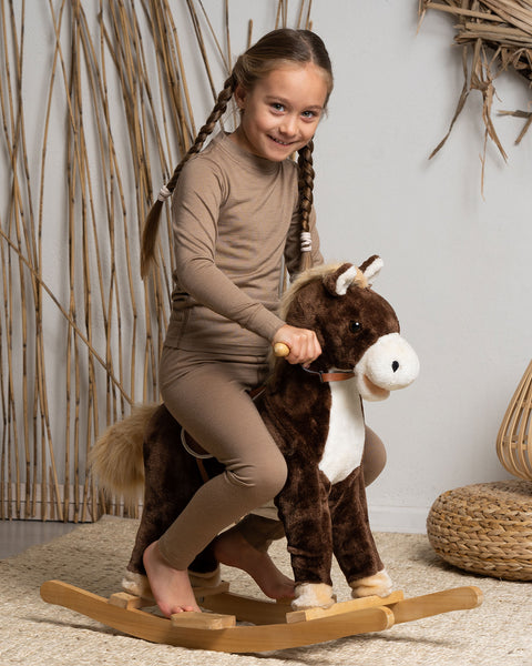 A smiling young girl with braided hair rides a plush brown and white rocking horse indoors. She is dressed comfortably in a matching tan long-sleeved top and leggings.