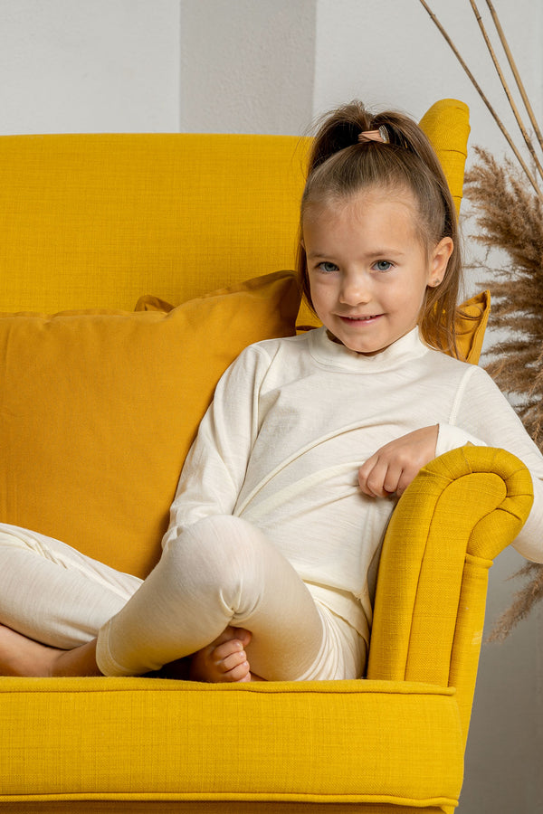 A girl sitting in a yellow armchair, with her legs crossed, wearing the natural color menique long sleeve merino 160gsm top in natural color. 