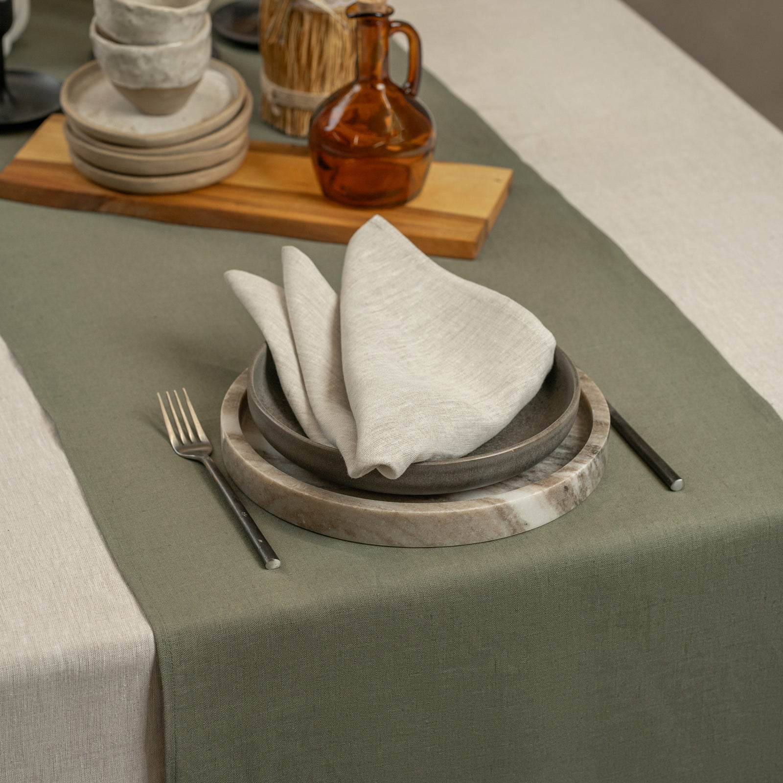 A close-up view of several folded, textured linen napkins in earthy tones like olive green, mustard yellow, rust red, and light beige, scattered on a white surface. The front two napkins are wrapped in brown kraft paper sleeves printed with the brand name "MENIQUE" and feature round brown stickers that read "MADE FROM 100% LINEN."