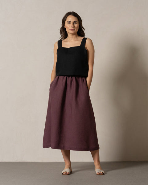 A woman with medium-toned skin and dark hair stands facing the camera against a neutral background. She is wearing a pure black, sleeveless square-neck linen top and a flowy, maxi skirt in a shadow purple color, with her hands casually placed in the skirt&#39;s pockets and light-colored sandals on her feet.