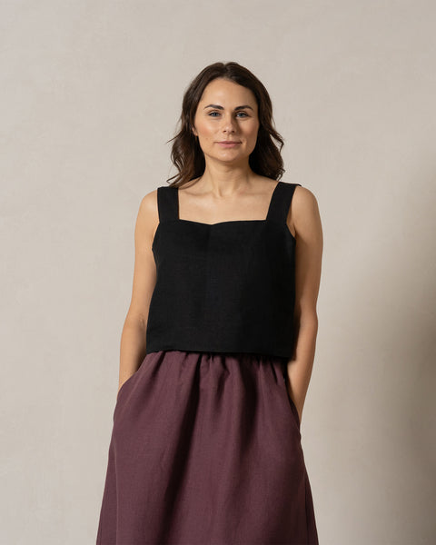 A woman is elegantly dressed in a pure black square-neck linen top and menique maxi skirt. The image captures her from the waist up, showcasing the flattering neckline. The lighting is soft and even, highlighting the texture and color of her clothing.