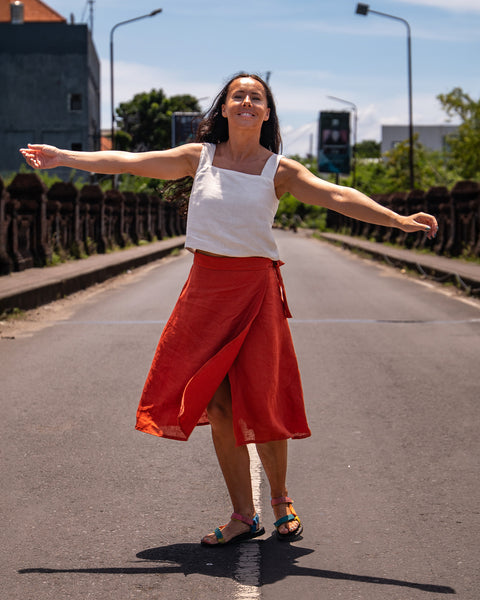 A woman with shoulder-length dark hair, wearing a pure white linen sleeveless crop top, menique linen wrap skirt, and sandals, stands with outstretched arms and a smile on a paved road under a bright sky.