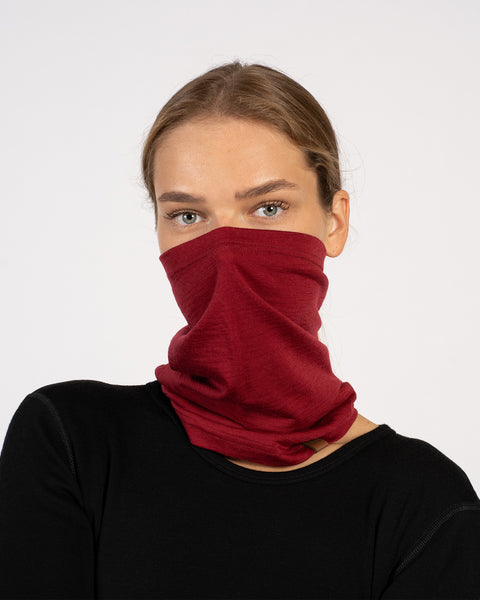 A studio shot of a woman looking directly at the camera against a plain white background. She is wearing a black long-sleeved top and a dark red neck gaiter that is pulled up to cover her lower face, nose, and neck.