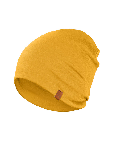 A close-up shot of a spicy yellow merino wool menique beanie hat is presented against a stark white background. The beanie has a slightly textured appearance and features a small, brown rectangular tag sewn onto its folded brim. The hat is softly draped and shows gentle wrinkles in the fabric.