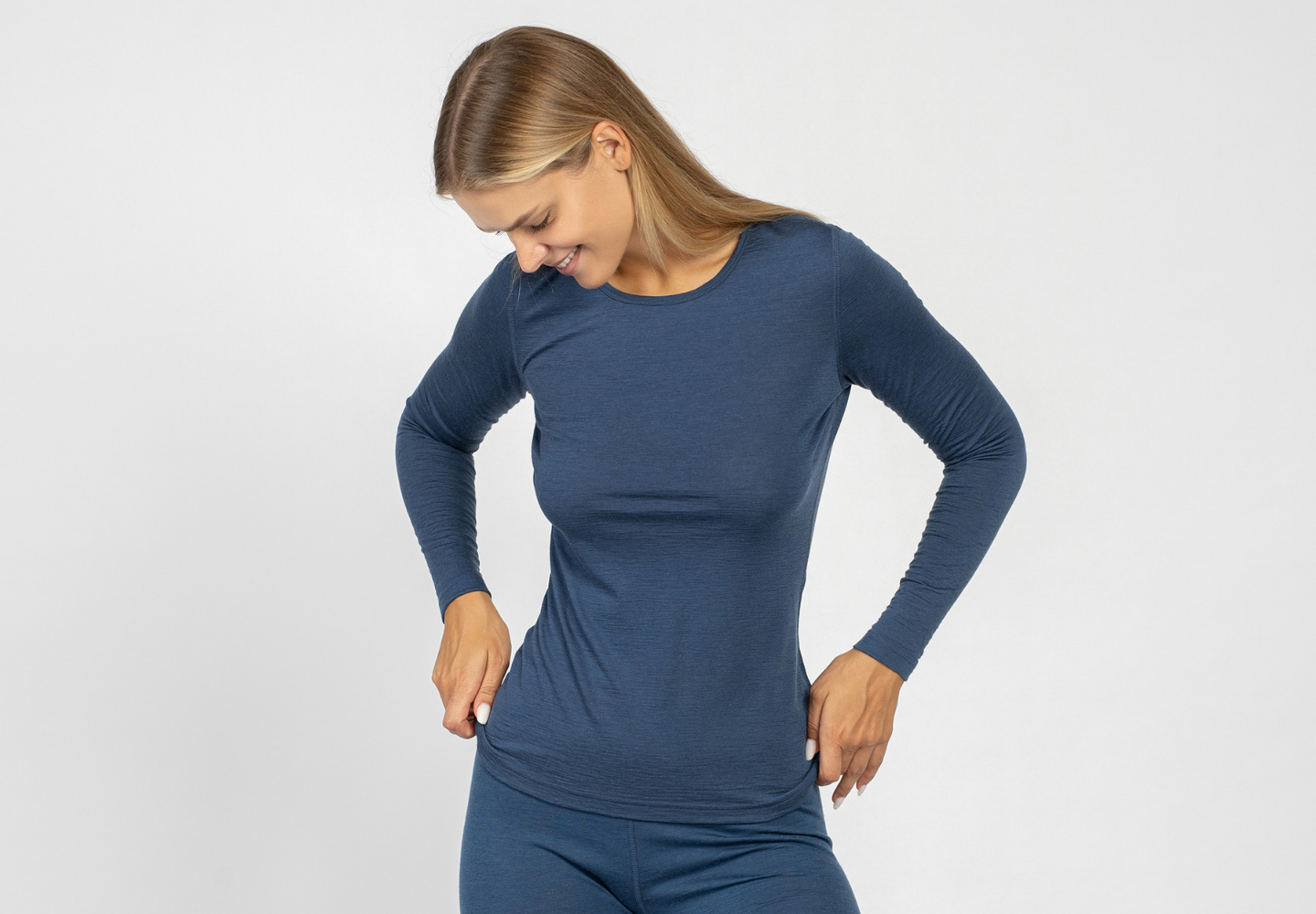 A smiling woman wearing a fitted long-sleeve navy merino wool top and matching bottoms from menique, looking down with her hands resting at her hips against a white background.